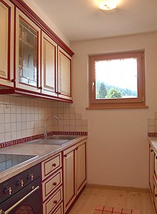 Appartamento a Soraga di Fassa. Il cucinino. E' dotato di cucina in vetroceramica, forno elettrico, lavastoviglie, frigorifero con capiente freezer. L'arredo è completamente in legno color beige con cornici rosse bordeaux.