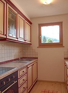 Appartamento a Soraga di Fassa. Il cucinino. E' dotato di cucina in vetroceramica, lavastoviglie, frigorifero con capiente freezer. L'arredo è completamente in legno color beige con cornici rosse bordeaux.